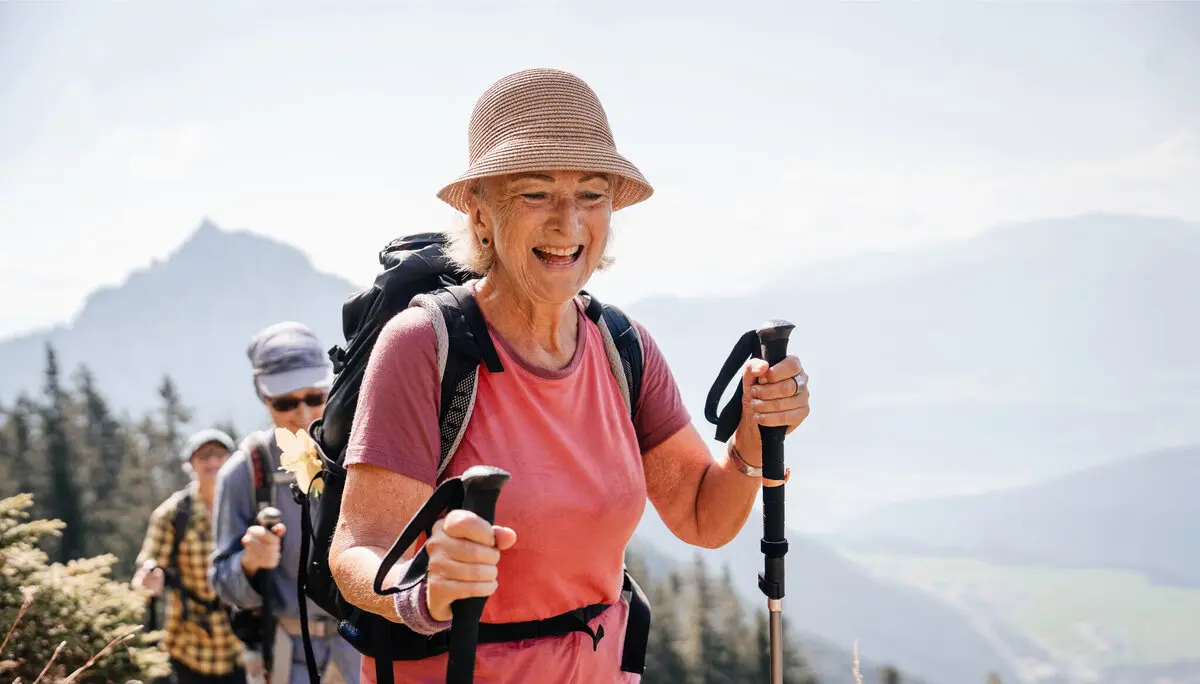 Eine ältere Frau mit Stöcken und Sonnenhut führt glücklich eine Wandergruppe an. | © DAV / Marisa Koch